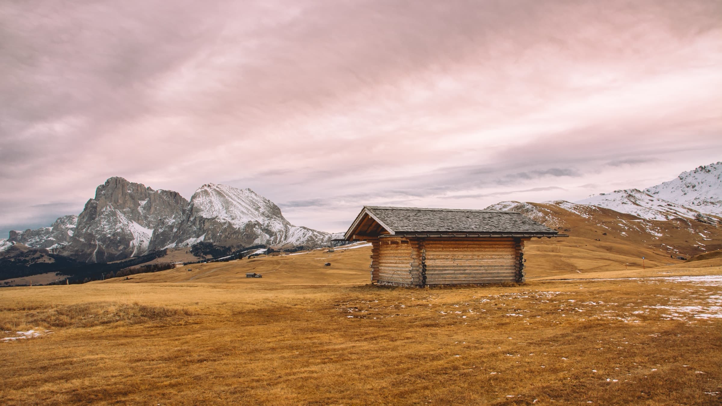 A lone wooden cabin on a golden meadow below snowy peaks and a pink sky